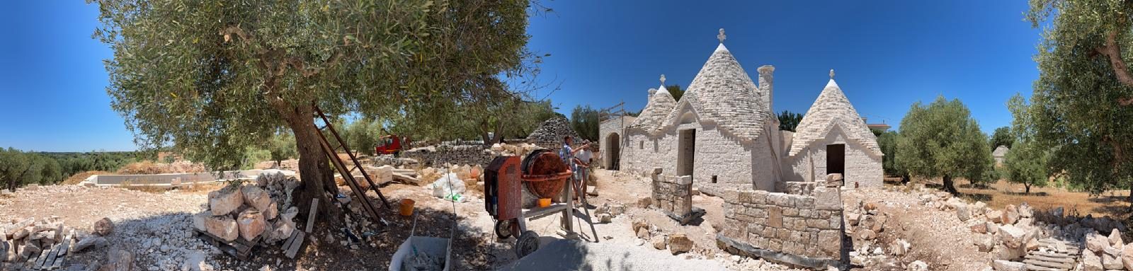 A wide panorama of the estate mid-restoration — cement mixer, gathered stone and the whitened trulli beneath the olive trees