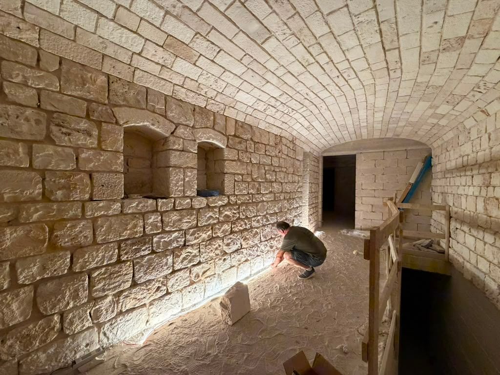A barrel-vaulted stone room, freshly pointed, with a mason working along the base of the wall