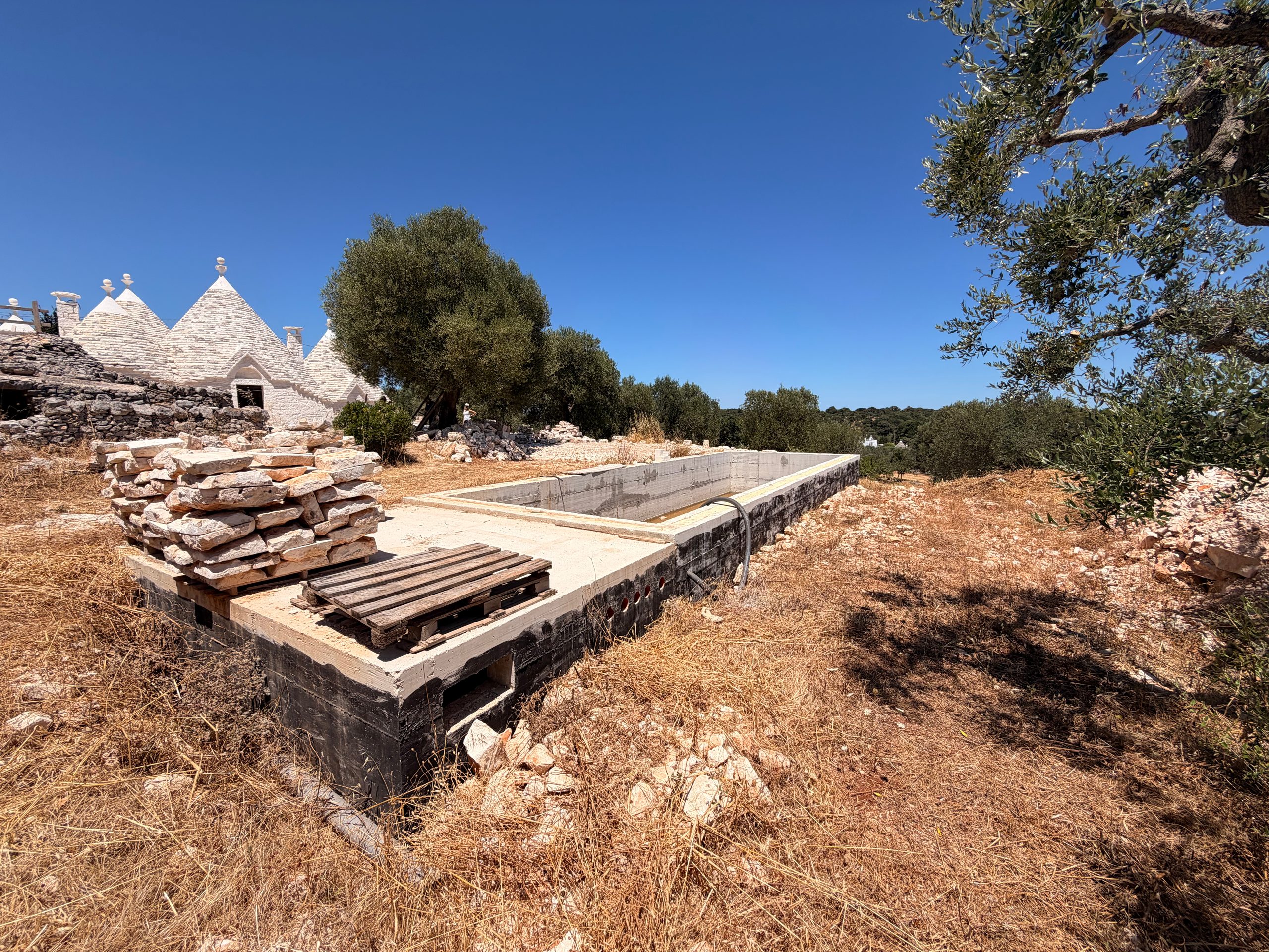 The concrete shell of the water tank set into the dry grass, the trulli standing beyond it